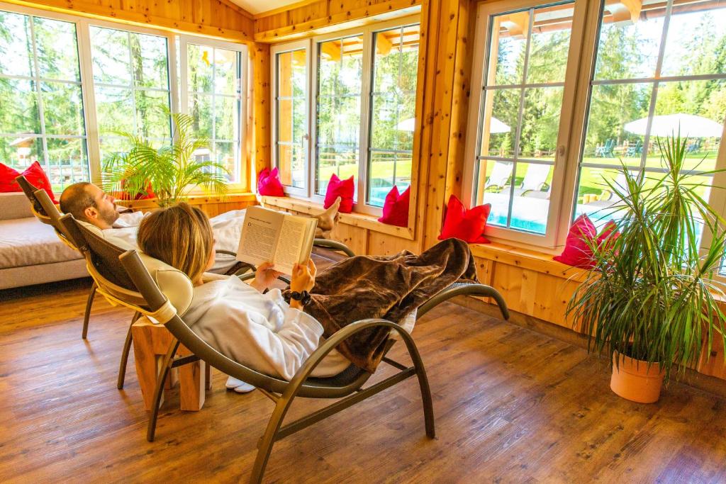 Relaksējoša telpa ar lieliem logiem un dīvānu, kur divi cilvēki atpūšas, viens lasa grāmatu, bet otrs relaksējas. Telpā ir zaļas augu rotājumi un spilgti spilveni.