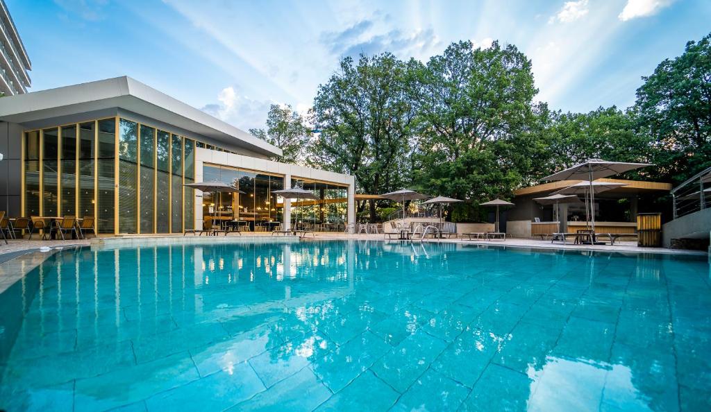 Luksusīgs baseins ar skatu uz modernu ēku un koku ainavu, kas nodrošina relaksējošu atmosfēru. Ideāla vieta atpūtai un izklaidei.