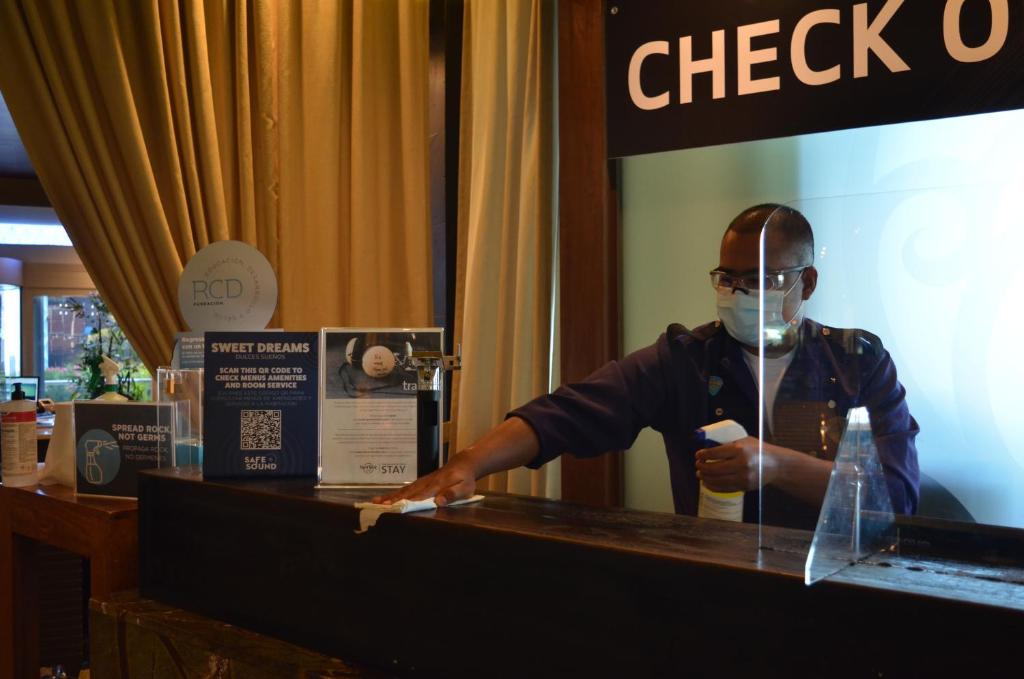 Reģistrācijas process pie Hard Rock Hotel Cancun ar viesmīli, kas dezinficē galdu.