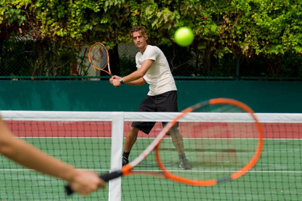 Tennisists sitien bumbiņu laukumā, gatavojoties izsist to atpakaļ. Fons ir zaļa siena ar augiem un citiem tenisistiem.