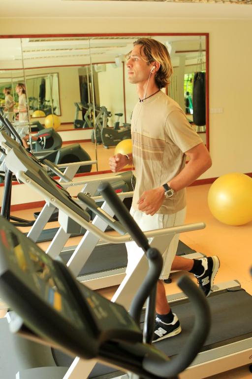 Vīrietis skrien uz trenažiera sporta zālē, klausoties mūziku austiņās. Aiz viņa redzami citi trenažieri un sporta aprīkojums.