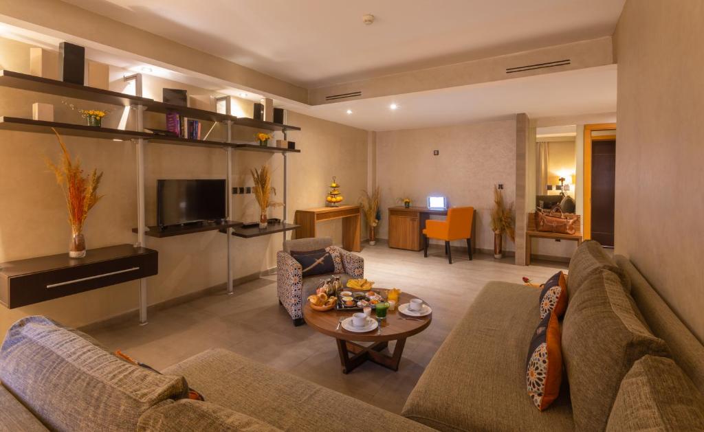 Mūsdienīgs viesnīcas numurs ar platām dīvāniem, televizoru un koka galdu, uz kura ir dažādi dzērieni un uzkodas. Telpā ir siltas krāsas un dekoratīvi elementi, kas rada mājīgu atmosfēru.