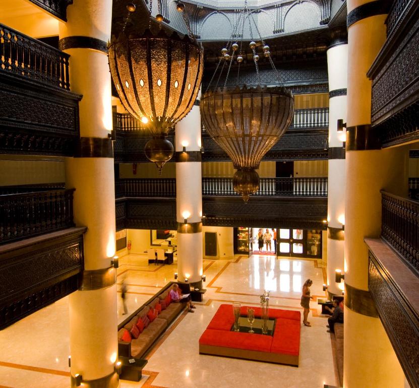 Lieliska viesnīcas halle ar elegantiem lustrām un modernu interjeru, kas rada greznu atmosfēru. Sarkanā dīvāna un dekoratīvo elementu izmantošana piešķir telpai siltumu un viesmīlību.
