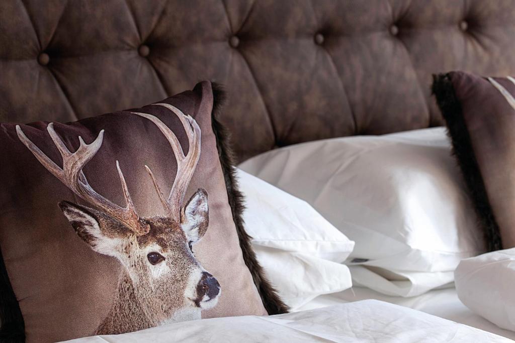 Dekoratīvs spilvens ar staltbrieža attēlu, novietots uz gultas ar baltām gultas veļām un tumši brūnu galvas klāju.