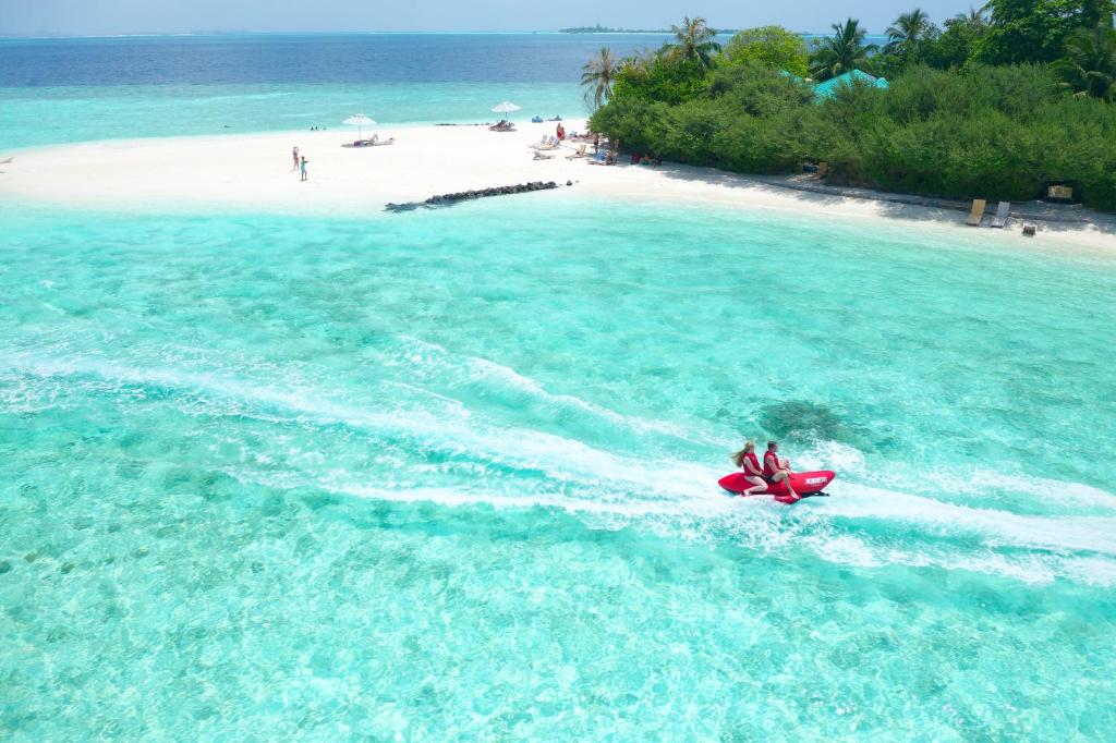 Divi cilvēki brauc ar ūdens motociklu pa skaidrām, tirkīza ūdeņiem, tuvumā baltā smilšu pludmale un zaļojoši koki. Ideāla tropu atpūtas ainava.
