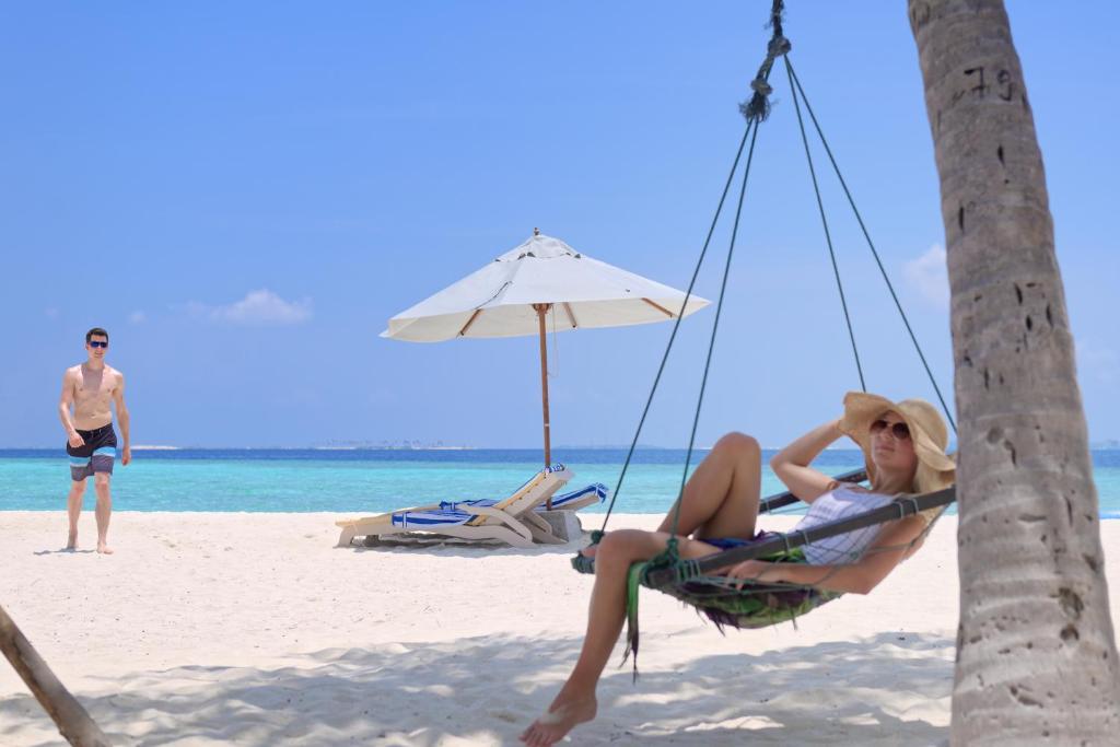 Pludmales ainava ar cilvēkiem, kas atpūšas zem saules, viena persona guļ šūpuļtīklā, bet otra pastaigājas pa smiltīm.