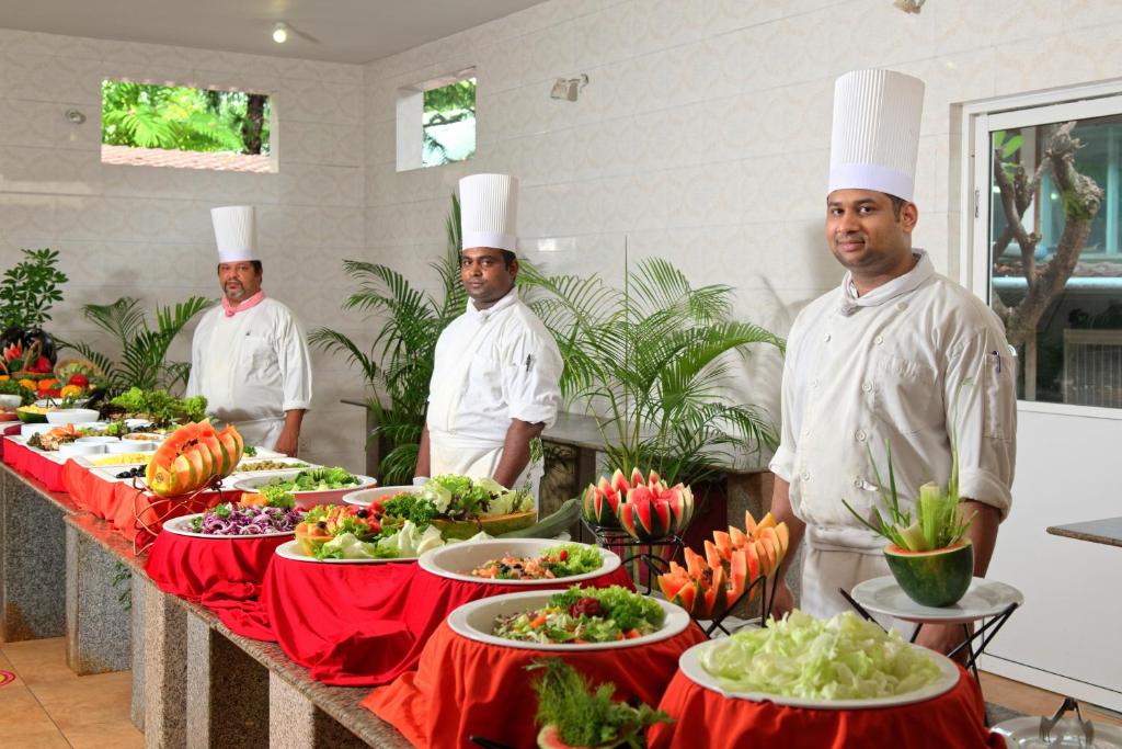 Šefpavāri stāv pie bagātīgas salātu un augļu bufetes, kas rotāta ar zaļumiem un krāsainām dekorācijām. Apskatāmi dažādi ēdieni uz galda, radot gardu un vilinošu atmosfēru.