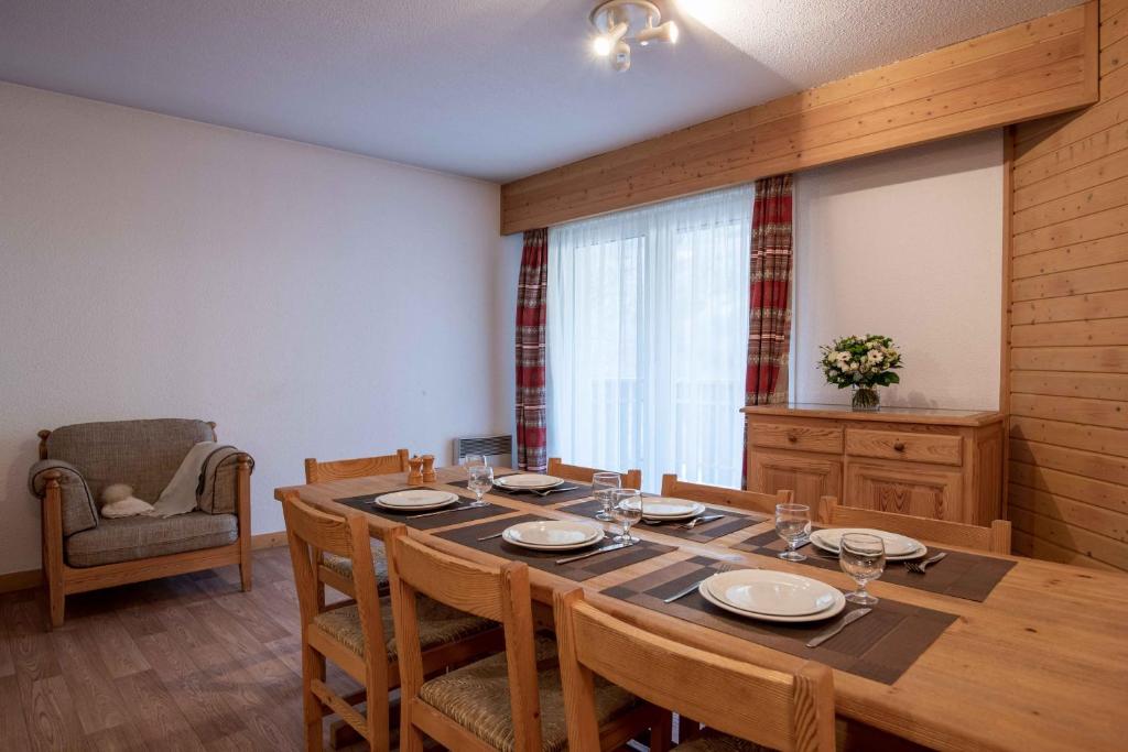 Mūsdienīga un mājīga ēdamistaba ar koka galdu un krēsliem, uzstādot galda piederumus un pušķi. Aizmugurē redzama gaismas caurspīdīga aizsarga, kas piešķir telpai gaišumu.