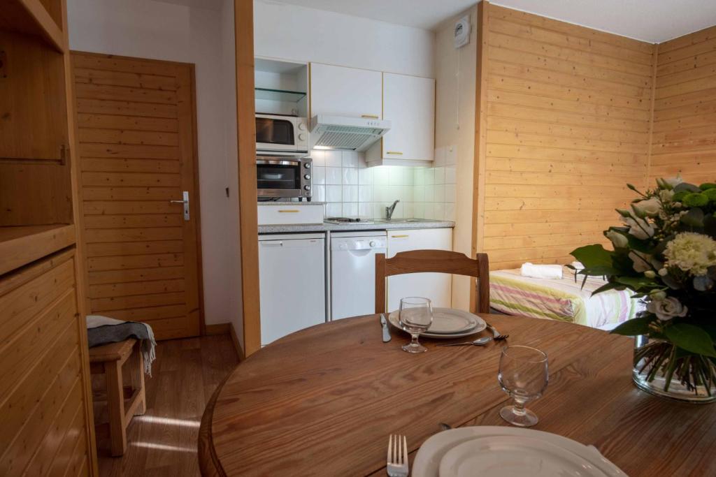 Mūsdienīga virtuve ar koka apdari un pusdienu galdu, kurā ir trauki un ziedu kompozīcija. Interjers ir ērts un viesmīlīgs, radot mājīgu atmosfēru.