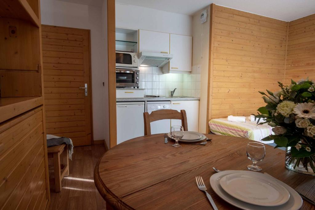 Mājīga virtuve ar koka sienām un galdautu, kas klāts ar galda piederumiem. Redzama modernā virtuve ar iekārtām, tostarp mikroviļņu krāsni un trauku mazgājamo mašīnu. Galdā ziedu kompozīcija, piešķirot telpai svaigumu.