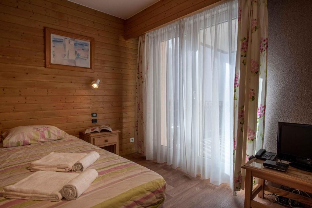 Mājīga guļamistaba ar koka sienām, gultu un logiem ar caurspīdīgām aizkariem. Telpā ir neliela koka galda un televizora stūra.