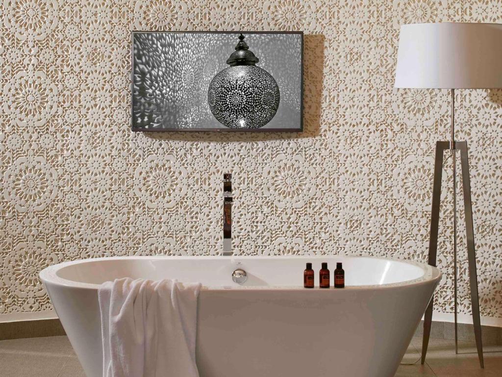 Mūsdienīgs vannas istabas interjers ar elegantu balto vannu, dekoratīvu sienu ar izgriezumiem un gaismas lampu. Uz vannas atrodas aromātiskās eļļas pudelītes, radot relaksējošu atmosfēru.