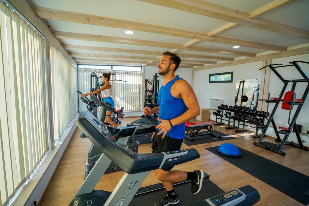 Divi cilvēki vingro sporta zālē, izmantojot kardio iekārtas un svarus. Gaismas telpa ar koka griestiem rada motivējošu atmosfēru treniņiem.