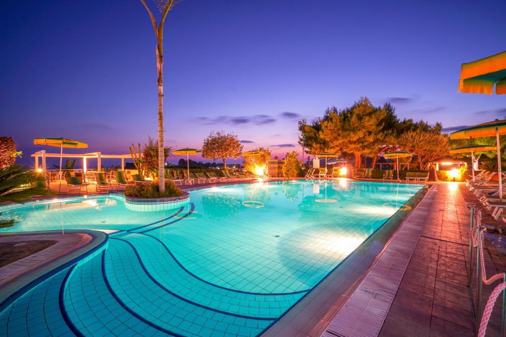 Peldbaseins ar zilu ūdeni un apgaismojumu, ieskauts ar zaļām kokiem un sauļošanās krēsliem, pie saulrieta. Ideāla vieta atpūtai un relaksācijai.