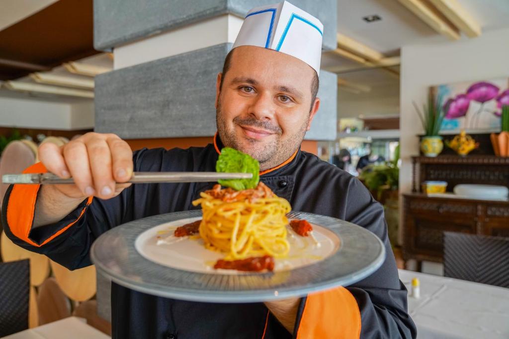 Šefpavārs lepni pasniedz šķīvi ar pasta ēdienu, rotātu ar zaļumiem un tomātiem, restorānā. Fons ietver elegantu interjeru ar dekoratīvām detaļām.