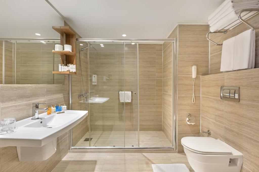 Mūsdienīgs vannas istabas interjers ar stikla dušu, izsmalcinātu izlietni un koka plauktu, kas satur higiēnas piederumus. Gaiši brūnas flīzes piešķir siltumu telpai.