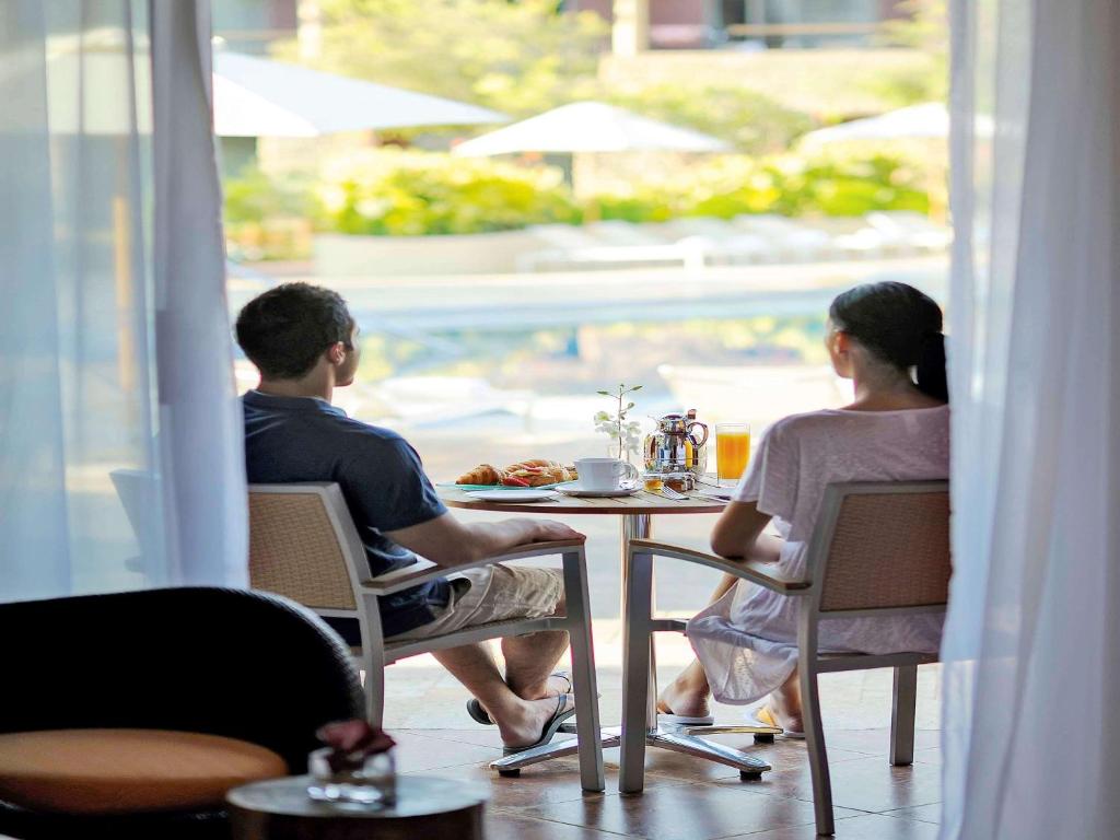 Pāris, kas sēž pie galda ar brokastīm, baudot skatu uz peldbaseinu un dārzu, caur caurspīdīgu aizkaru.