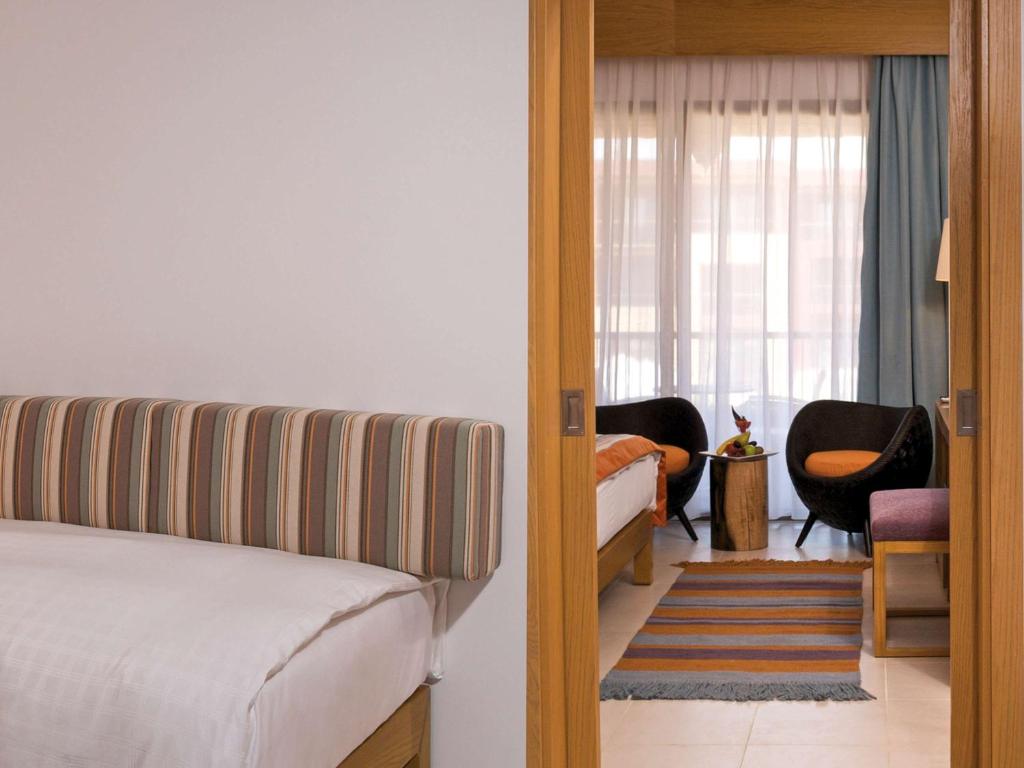 Mūsdienīga viesnīcas istaba ar stilīgu gultu un ērtu atpūtas zonu, kurā redzami melni krēsli un dekoratīvs paklājs.