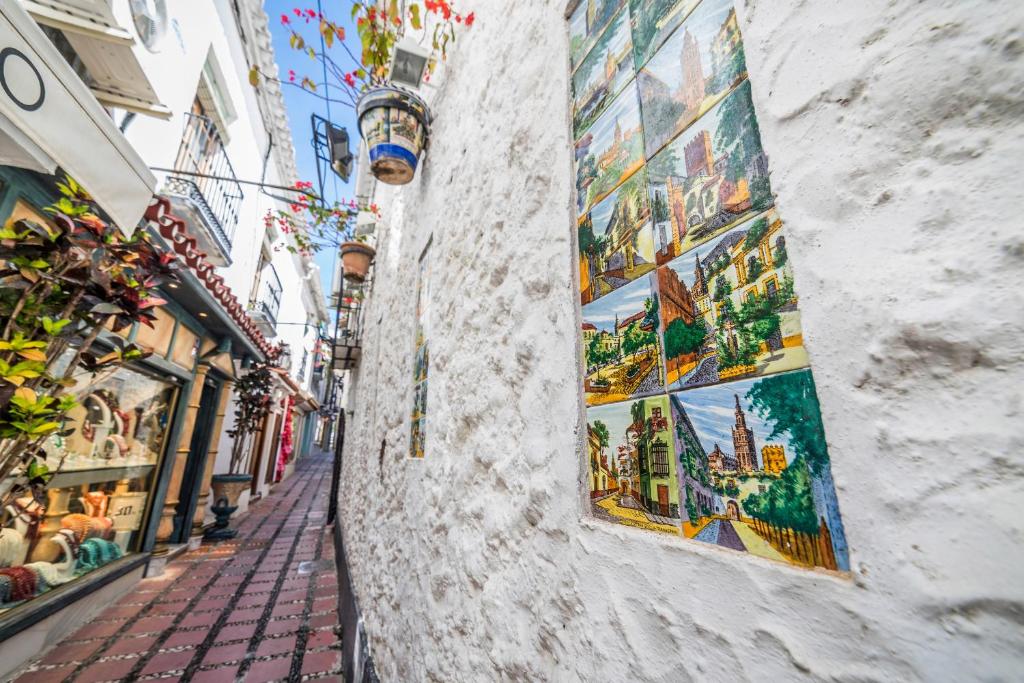 Šaurs ieliņš ar balto mūri, kurā ir krāsaini mākslas darbi, un ziedoši augi, kas karājas no jumta. Apkārt ir veikali ar vietējiem izstrādājumiem.