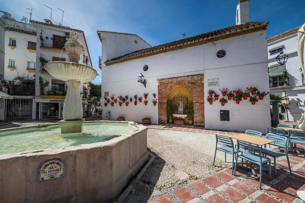 Burvīgs laukums ar strūklaku un ziedu podiņiem pie baltas ēkas, kurā ir arkas ieeja. Apkārtējās kafejnīcas piedāvā patīkamu atpūtu saulainā dienā.