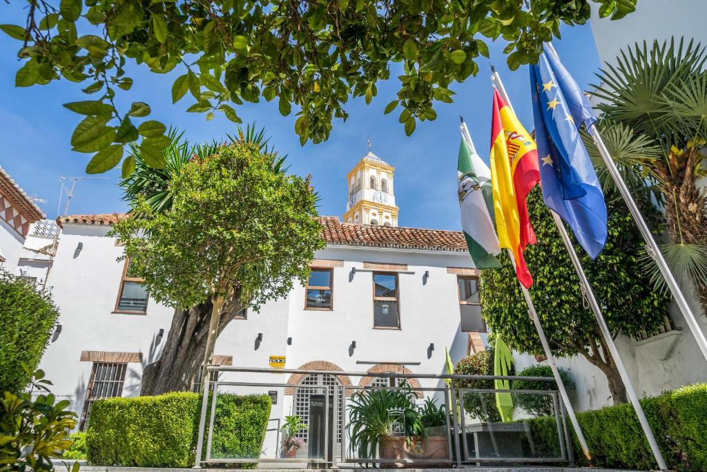 Vēsturiska ēka ar zaļām augu kompozīcijām un dažādām karogiem priekšplānā, fons ir skaidra debess ar torni.