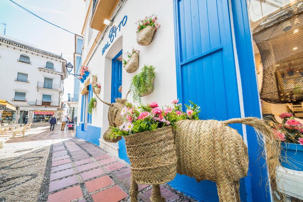 Bļodā izveidots zālītes dzīvnieks ar ziediem, kas rotā zilo ēku šaurā ielā. Apkārt esošās mājas un cilvēki dod pilsētai dzīvīgumu.