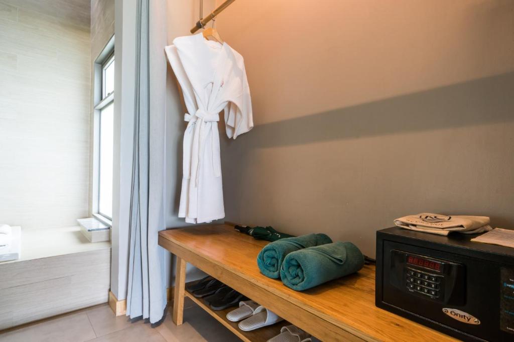 Elegants vannas istabas stūris ar baltu peldmēteli, diviem zaļiem dvieļiem un seifu uz koka galda. Gaismas tonis ir gaišs un mūsdienīgs, radot relaksējošu atmosfēru.