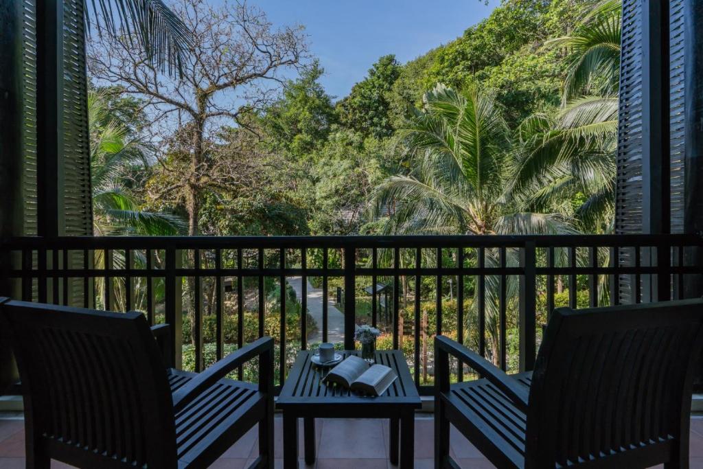 Balkons ar divām krēslām un skatu uz zaļu dārzu, kas apņem palmām un kokiem. Uz galda atrodas grāmata un krūze.