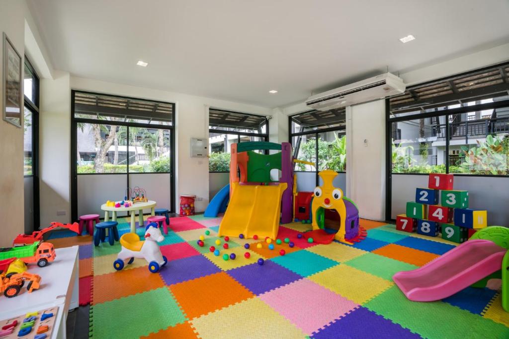 Krāsains bērnu rotaļu istaba ar dažādiem rotaļlietu elementiem, tostarp slidkalniņu, rotaļu māju un krāsainām mīkstajām paklāju flīzēm. Telpa ir gaiša ar logiem, kas sniedz skatu uz dārzu.