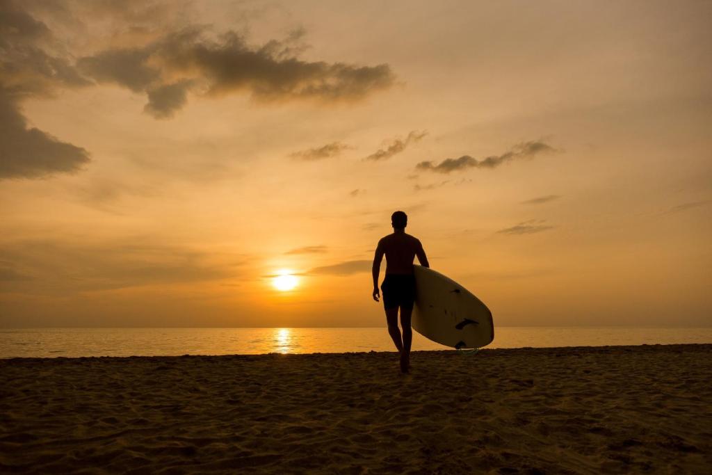 Sērfošanas entuziasts iet pa pludmali saulrieta gaismā, nesot sērfošanas dēli. Aiz viņa redzama mierīga jūra un krāšņa debesis ar zeltainiem toņiem.