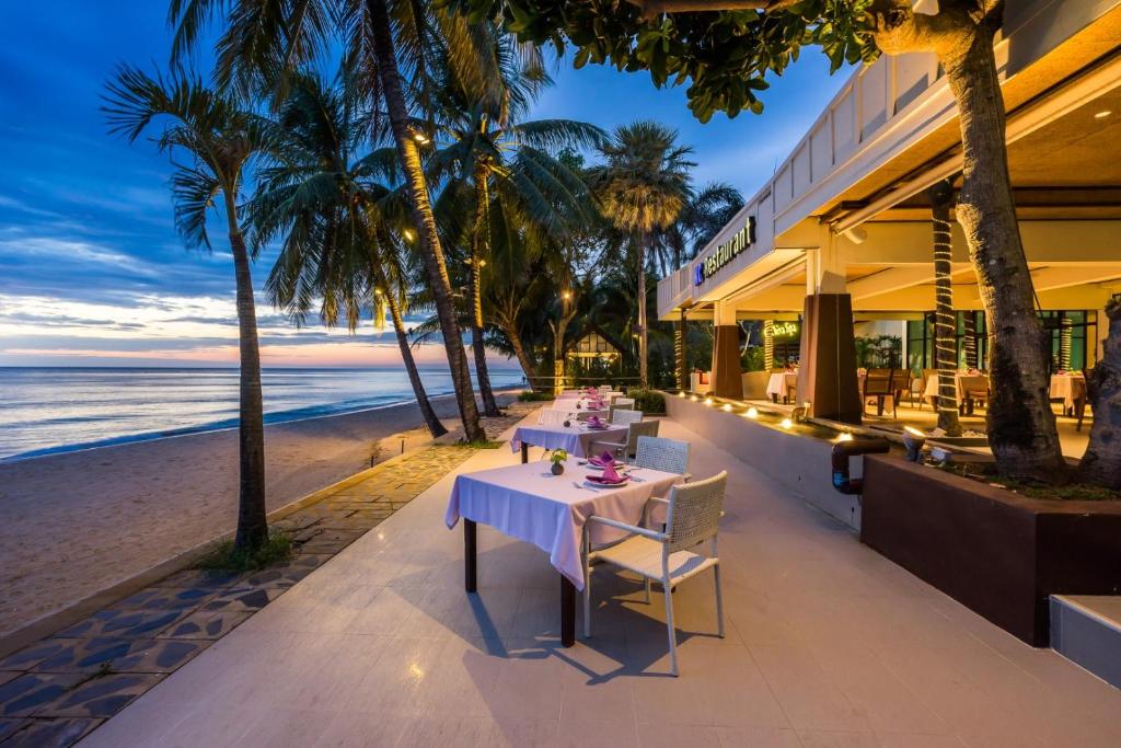 Pludmales restorāna terase ar galdiem, kas saklāti romantiskai vakariņai, apņemta ar palmām un blakus jūrai. Saulrieta gaisma rada patīkamu atmosfēru.