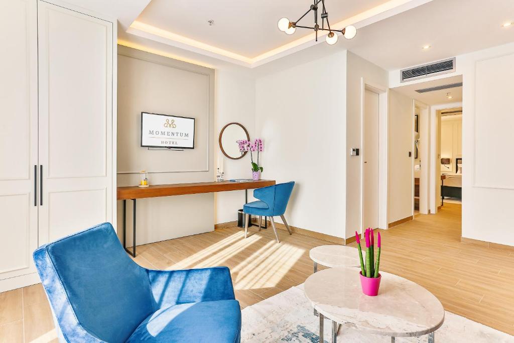 Mūsdienīgs viesnīcas numurs ar stilīgu interjeru, iekārtots ar zilu krēslu, koka galdu un ziediem. Redzama viesnīcas zīme "Momentum Hotel".