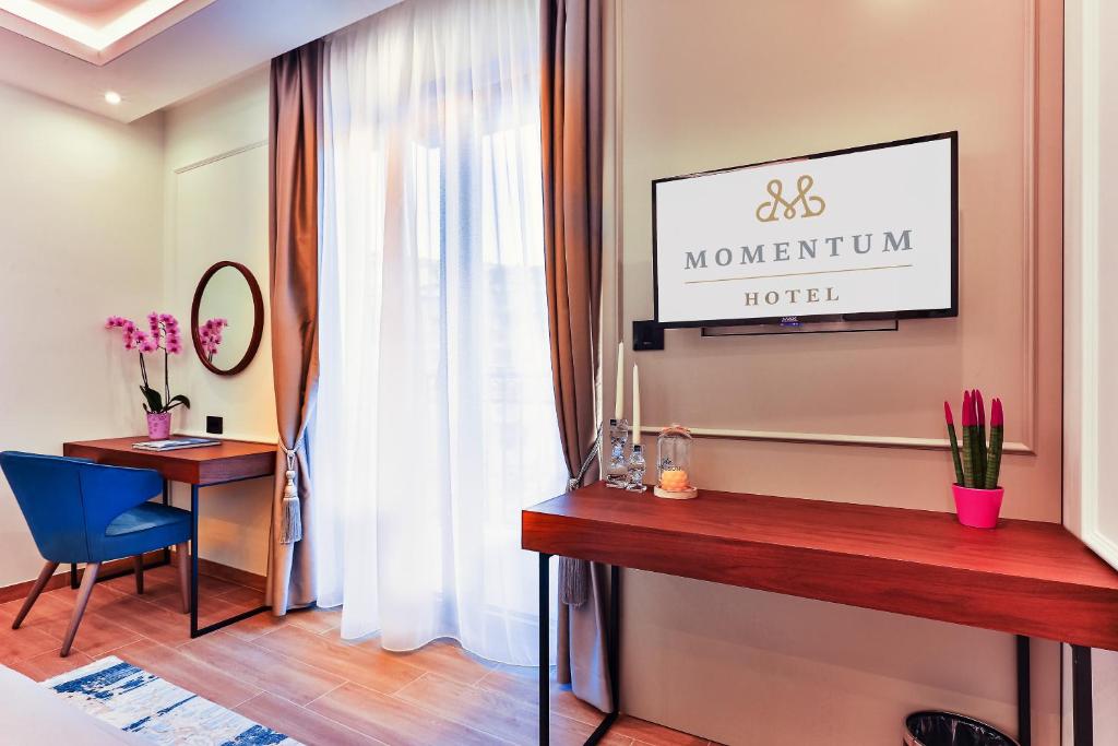 Mūsdienīga viesnīcas istaba ar koka mēbelēm, ziliem krēsliem un dekoratīvām orhidejām. Uz sienas redzams "Momentum Hotel" logotips.