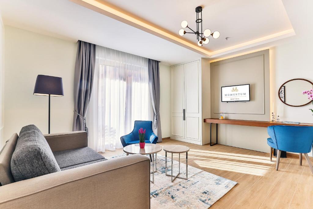 Mūsdienīgs viesnīcas numurs ar ērtu dīvānu, krāsainiem krēsliem un koka galdu, kas nodrošina patīkamu atmosfēru. Interjeru papildina dabīga gaisma un dekoratīvi elementi.