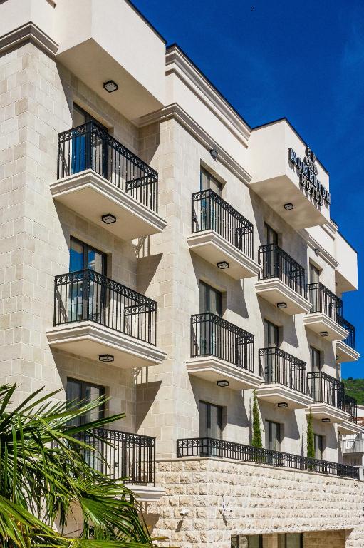 Mūsdienīga viesnīca ar balkoniem un elegantu arhitektūru, izvietota zem spožas debess. Šis attēls izceļ ēkas stilīgo dizainu un apkārtējo zaļumu.