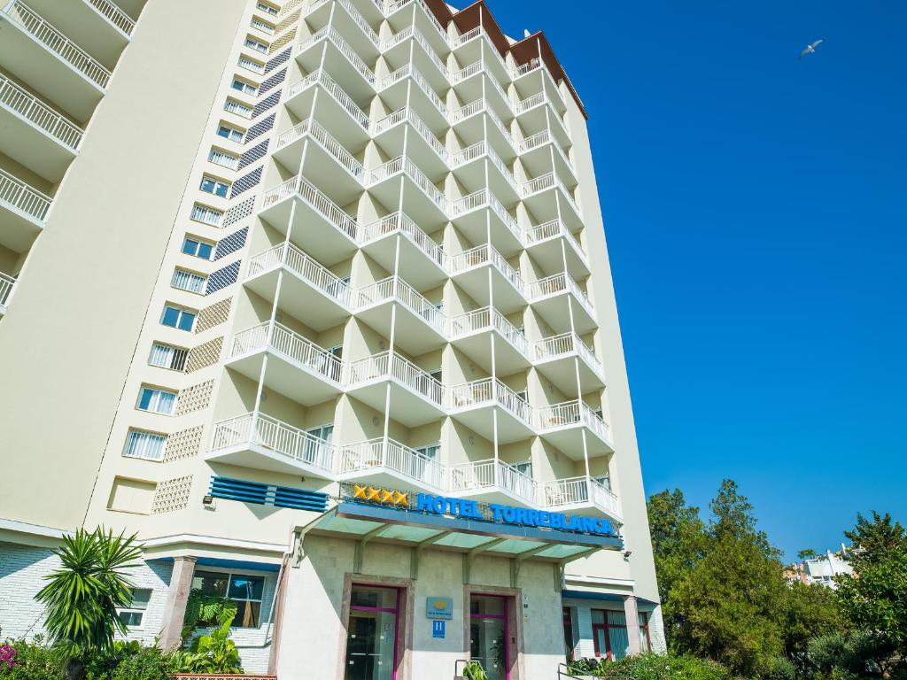 Moderns viesnīcas ēkas eksterjers ar plašām terasēm un skaidru zilo debesu fonu. Viesnīcas nosaukums "Hotel Torreblanca" ir redzams virs ieejas.