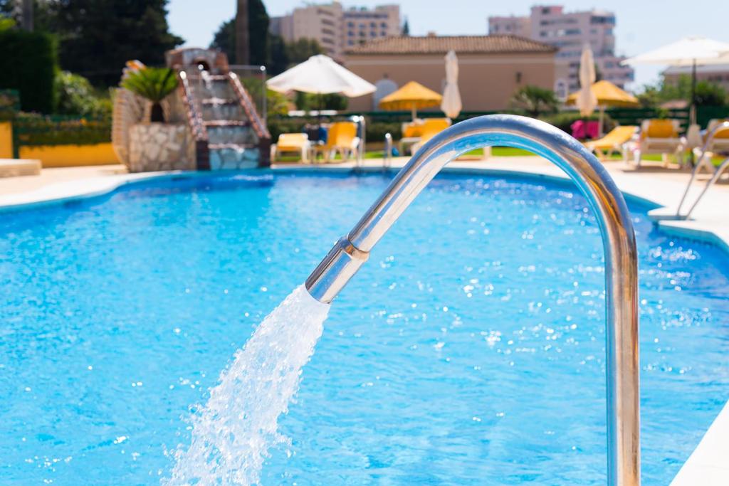 Peldbaseins ar spožu zilu ūdeni, no stieples kāpņu margas plūst ūdens. Apkārt baseinam redzami sauļošanās krēsli un saulessargi, radot relaksējošu atmosfēru.