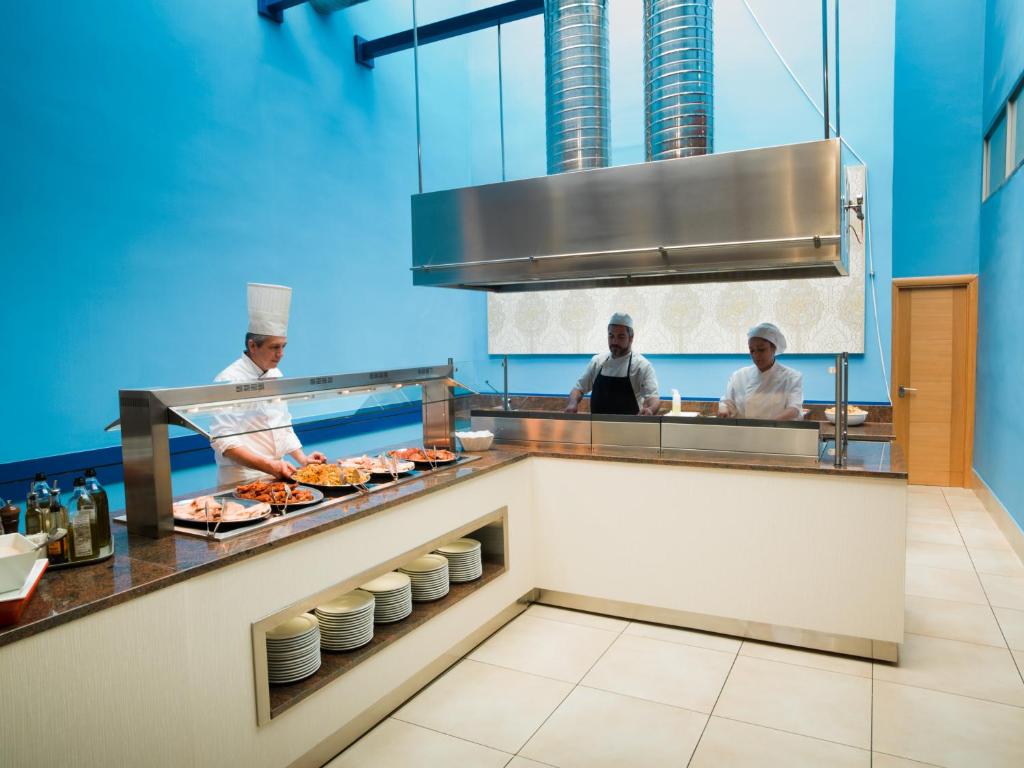 Šefpavārs gatavo ēdienus modernā virtuvē ar zilu apdari, kamēr divi palīgi strādā pie darba galda. Uz galda ir izlikti dažādi ēdieni, un virtuvē valda sakārtotība un profesionāla atmosfēra.