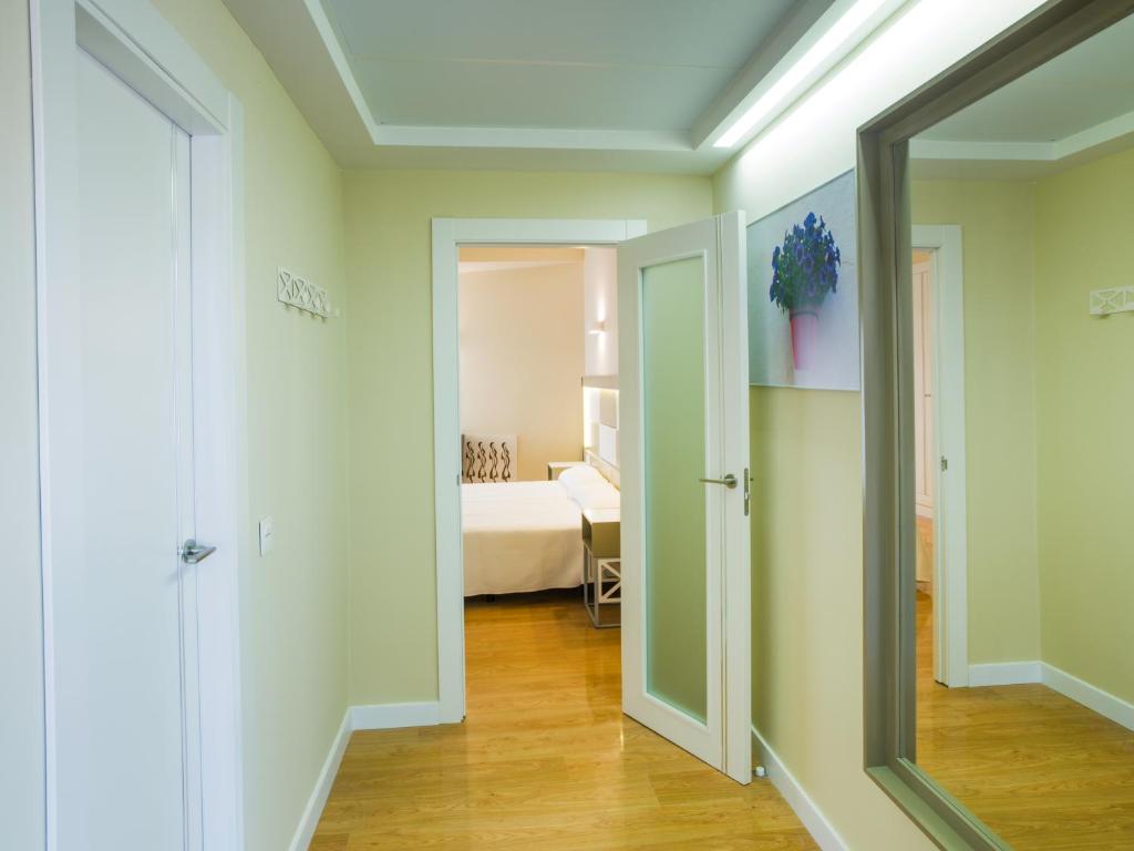 Gaiša un moderni iekārtota koridora ainava ar durvīm, kas ved uz guļamistabu. Redzama liela spoguļa un ziedu attēls uz sienas, radot mājīgu atmosfēru.