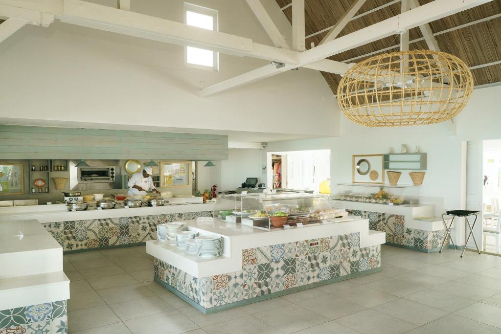 Mūsdienīga virtuve ar gaišu interjeru un stilīgu groza lampu, kur šefpavārs gatavo ēdienus. Uz galda redzami dažādi trauki un svaigi produkti.