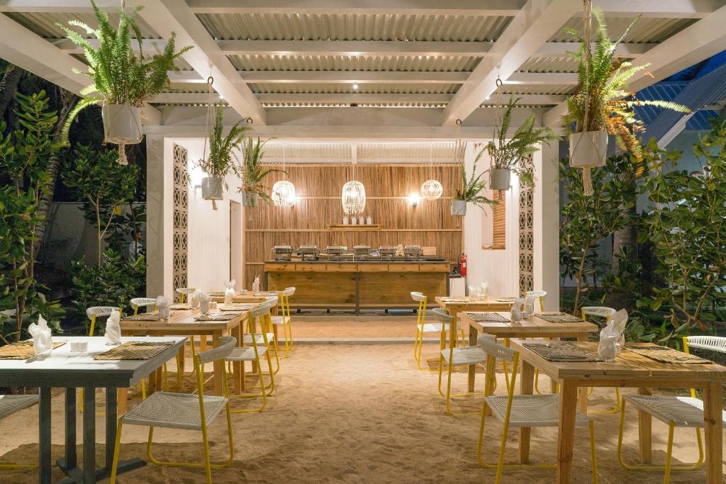 Moderns ārējais restorāns ar koka mēbelēm, spilgti dzeltenām krēsliem un zaļām augu kompozīcijām. Siltā apgaismojumā ir redzamas ēdamgaldi un bufetes zona.