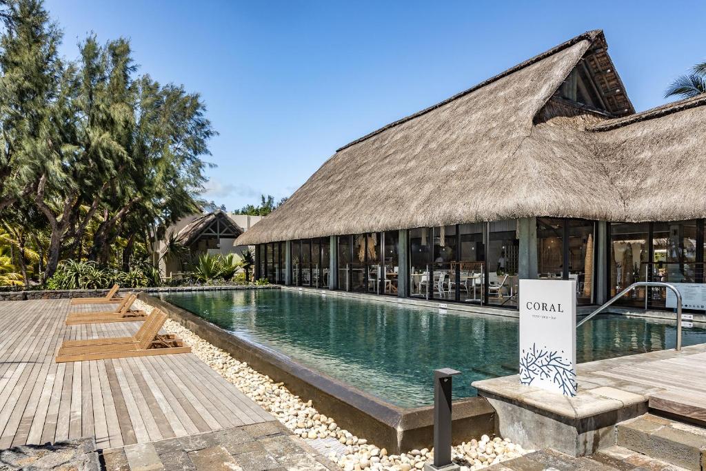 Viesnīcas komplekss ar baseinu un koka terasi, apzaļumots ar palmām un citām augiem, radot tropisku atmosfēru. Redzama ēka ar salmu jumtu un lieliem logiem.