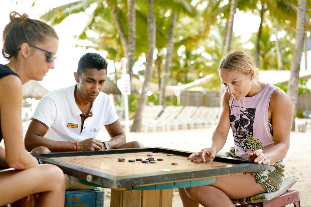 Divi cilvēki un vīrietis spēlē galda spēli tropiskā pludmalē, apkārt aug koki un saule spīd.