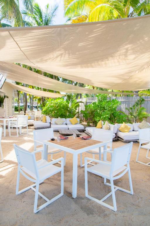 Brīvdabas kafejnīca ar baltām mēbelēm un dārzā izvietotām dīvānu vietām, kas ir ēnainas zem baltiem audumiem. Apkārt ir tropu augi un saulains laiks.