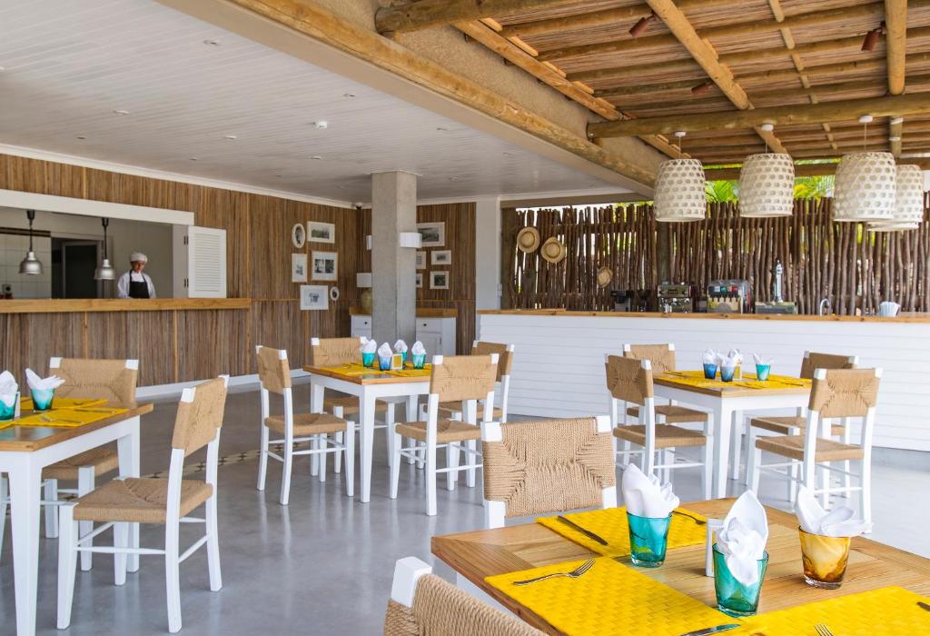 Mūsdienīgs restorāns ar gaišu interjeru, koka elementiem un krāsainiem galda piederumiem, kas rada viesmīlīgu atmosfēru. Redzams pavārs, kas strādā virtuvē.