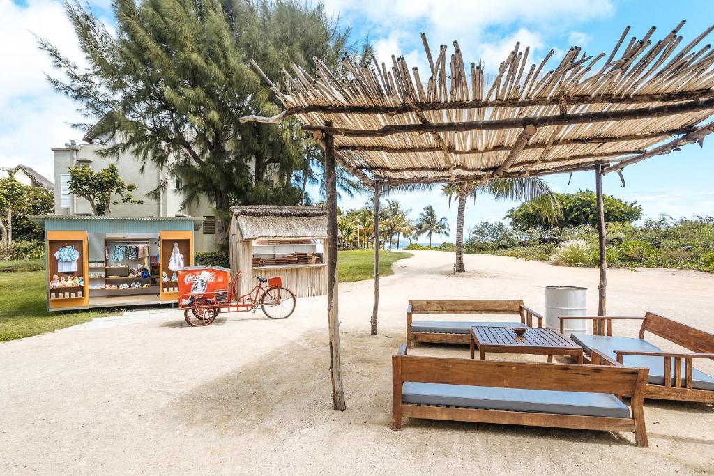 Pludmales atpūtas zona ar koka mēbelēm, saulessargu un tirdziņu, kurā redzama sarkanā Coca-Cola ratiņi. Apkārt aug zaļi koki un zāle, radot relaksējošu atmosfēru.