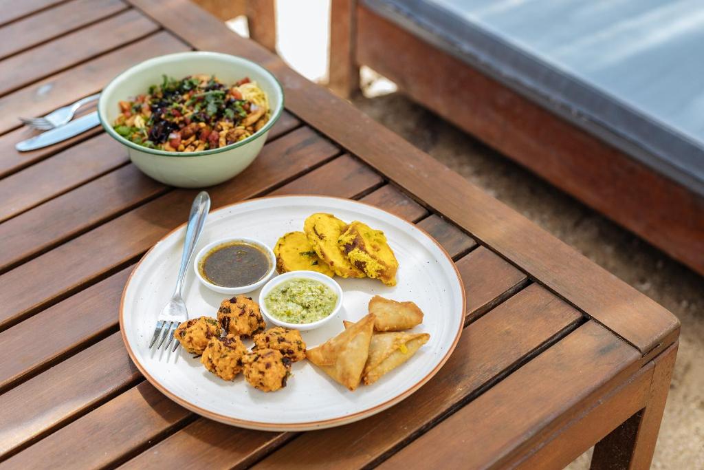 Svaigas maltītes plate ar ceptiem dārzeņiem, samosas un mērču trauciņiem, blakus zaļā salātu bļoda.