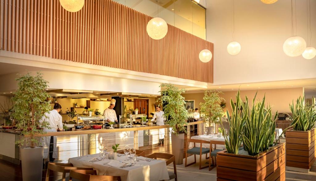 Mūsdienīgs restorāns ar atvērtu virtuvi un zaļiem augiem dekorā, kur šefpavāri gatavo ēdienus. Gaisotni rada eleganti gaismas ķermeņi un koka akcenti.