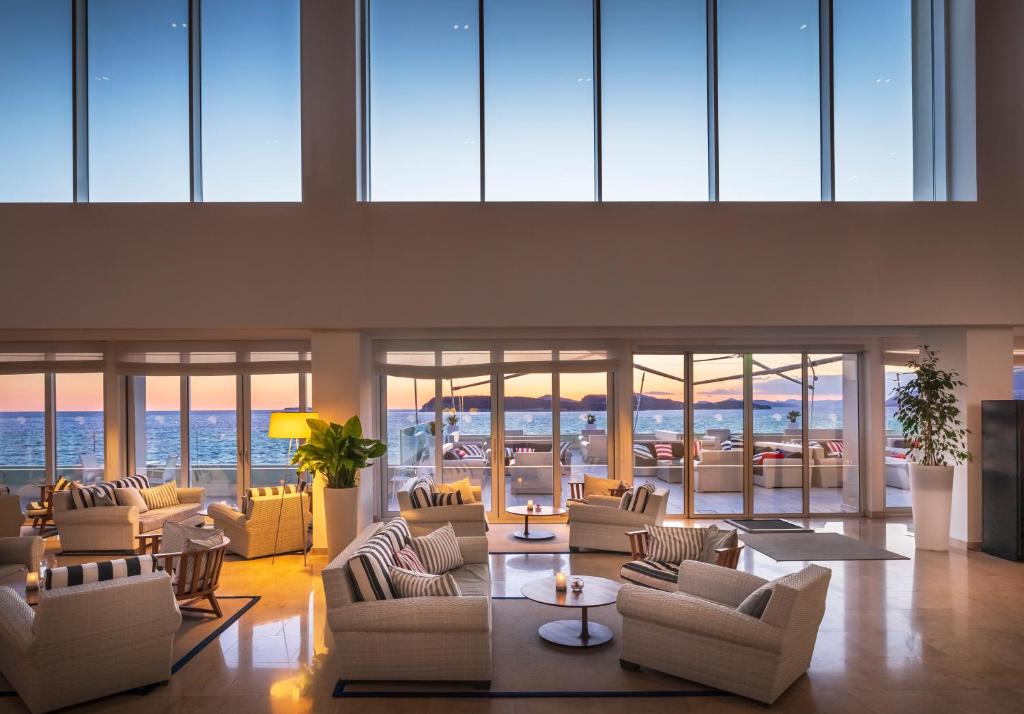 Mūsdienīgs viesnīcas interjers ar plašām logu sienām, caur kurām redzams jūras skats un krāsains saulriets. Atpūtas zona ir iekārtota ar ērtām krēsliem un galdiem, radot relaksējošu atmosfēru.