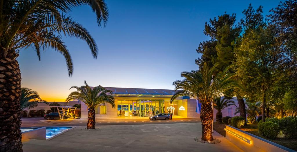 Moderns viesnīcas ieejas skats ar palmu kokiem un saulrieta gaismu. Viesnīcas arhitektūra ir mūsdienīga un pievilcīga.